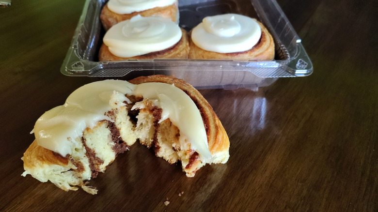 The striped cinnamon interior of a Whole Foods Market cinnamon roll is shown with three more in the background in a plastic clamshell topped with a thick swirl of frosting.