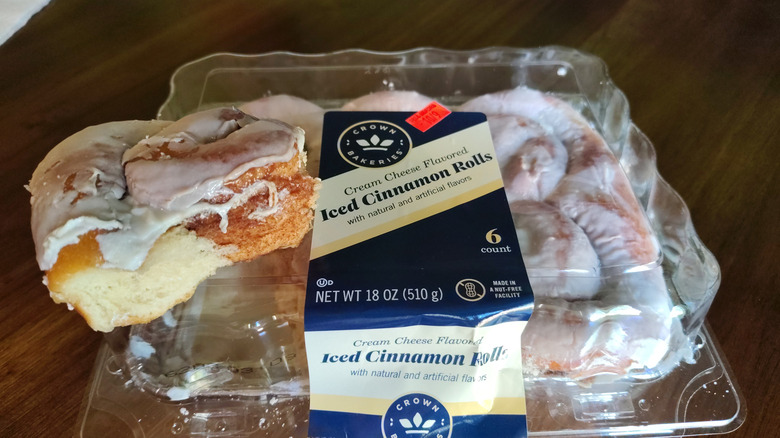 A Crown Bakery Cream Cheese flavored, iced cinnamon roll sits on top of a plastic clamshell full of cinnamon rolls.
