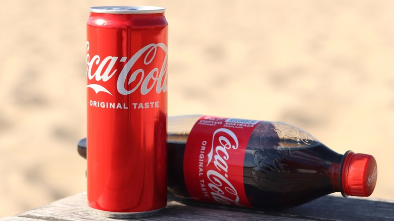 Can and bottle of Coca-Cola on a wood table outside.