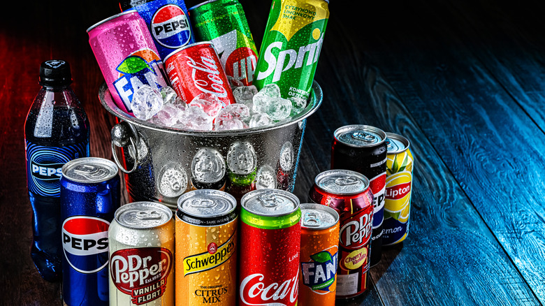 A bucket of ice filled and surrounded by various soda cans and bottles.
