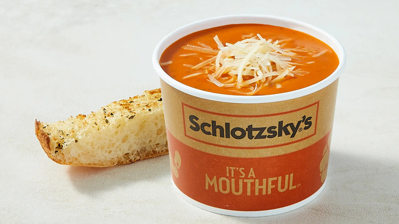 A paper bowl of Schlotzsky's Deli's Tomato Basil Soup topped with grated parmesan and a piece of bread