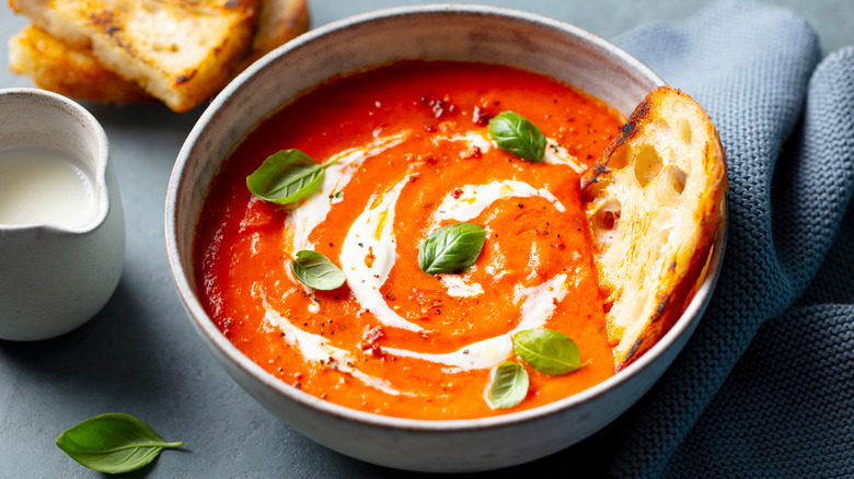 A bowl of tomato soup topped with cream and basil, and paired with a piece of bread