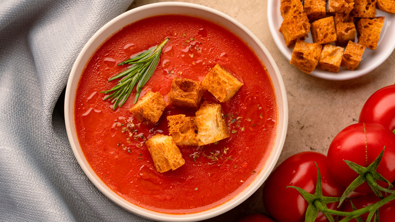 A bowl of tomato soup topped with croutons and rosemary with tomatoes next to it