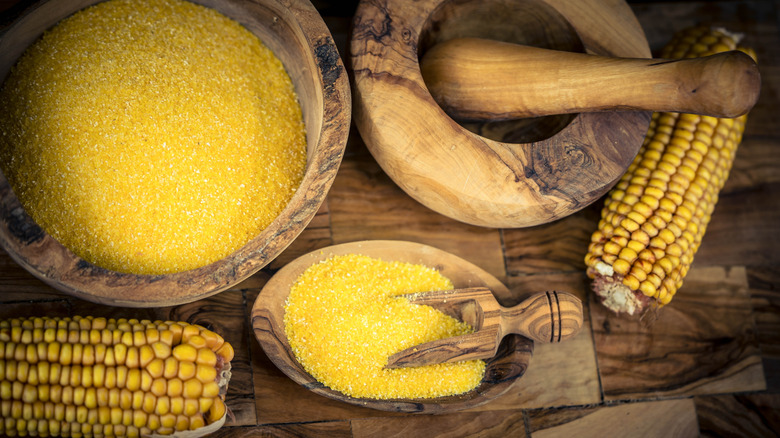 Wooden bowls full of cornmeal rest on a wooden table, surrounded by corn husks. One bowl has a small scoop in it.