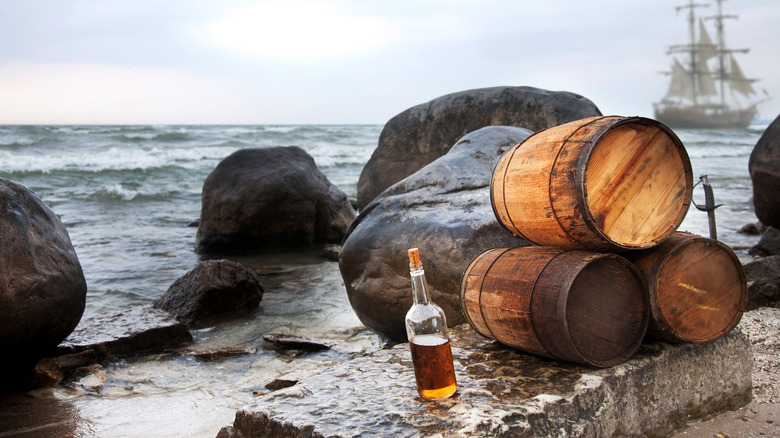 A corked bottle of rum sits to the left of a stack of wooden caskets at the edge of the ocean with wooden ship in background.