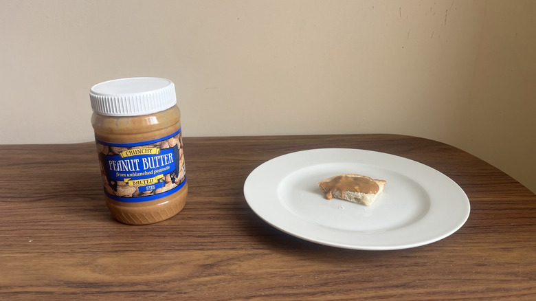 A jar of Trader Joe's crunchy salted peanut butter and some spread on a piece of bread on a round white plate