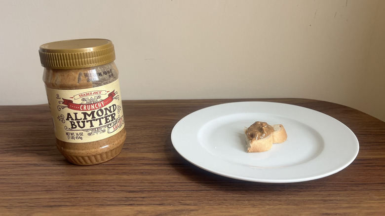 A jar of Trader Joe's crunchy salted almond butter and some spread on a piece of bread on a round white plate