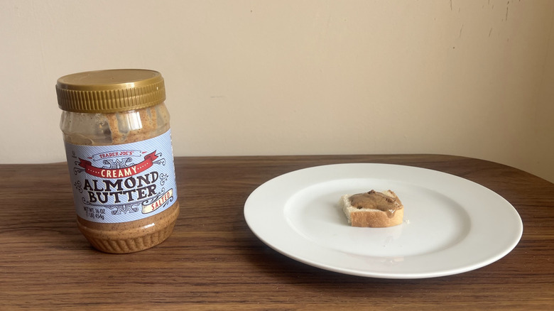 A jar of Trader Joe's creamy salted almond butter and some spread on a piece of bread on a round white plate