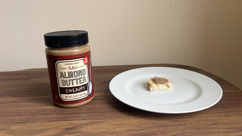 A jar of Trader Joe's raw almond butter and some spread on a piece of bread on a round white plate