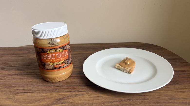 A jar of Trader Joe's creamy salted peanut butter and some spread on a piece of bread on a round white plate