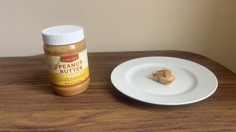 A jar of Trader Joe's peanut butter with honey and some spread on a small piece of bread on a round white plate
