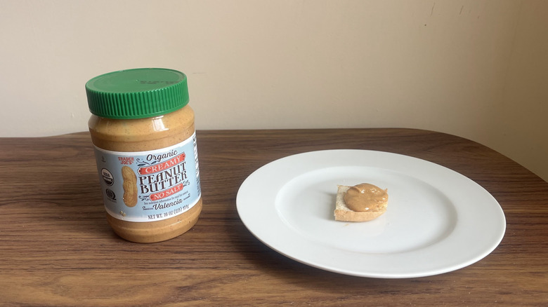 A jar of creamy organic peanut butter from Trader Joe's with some spread on a piece of bread on a round white plate