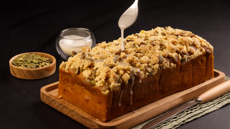 A loaf of pumpkin bread topped with a peanut butter glaze, pepitas, and streusel on a wooden cutting board