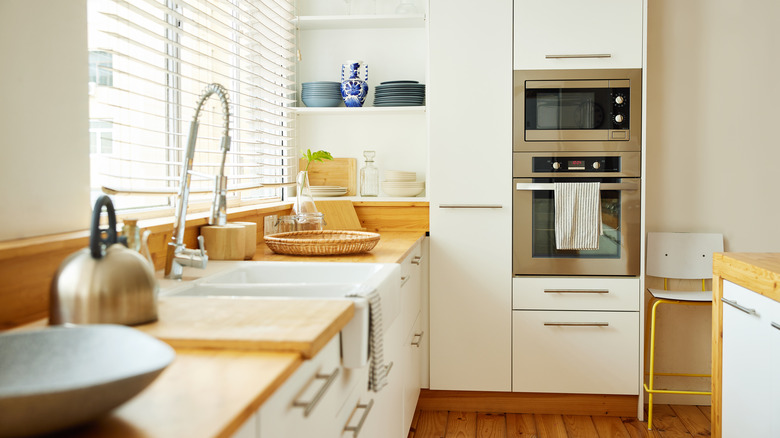 Plates and and drinking glasses arranged on shelf by oven, while a sink with faucet is on kitchen counter by window.