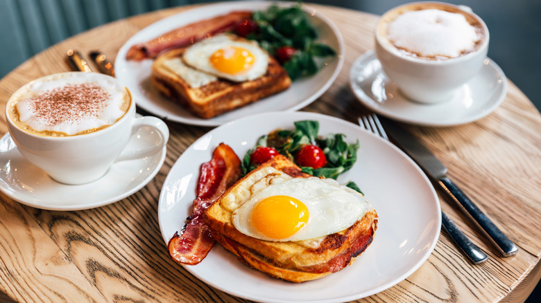 Two breakfast plates on a wooden table with fried eggs, toast, bacon, and side salad, with two lattes