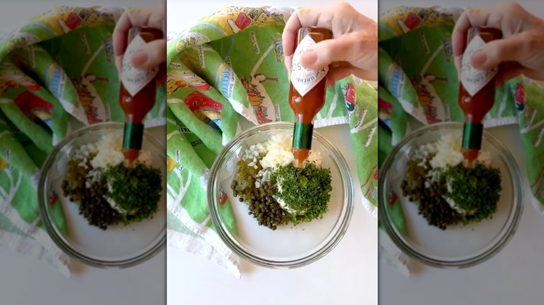 Hand pouring Tobasco sauce over glass bowl of tartar sauce ingredients