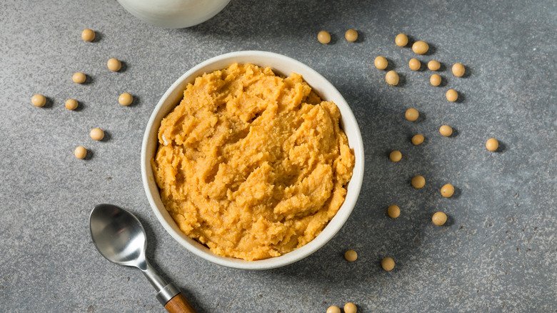 White miso paste in white bowl with soy beans scattered around and a metal spoon