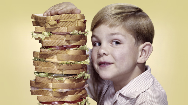 A young boy with a giant, multi-tiered sandwich dressed like he's from the 1950s.