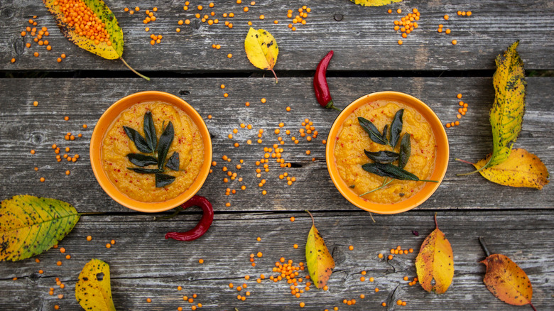 Fried sage leaves used to garnish two bowls of squash soup which are sitting on a wooden surface covered with autumn leaves, seeds, and a few random chile peppers