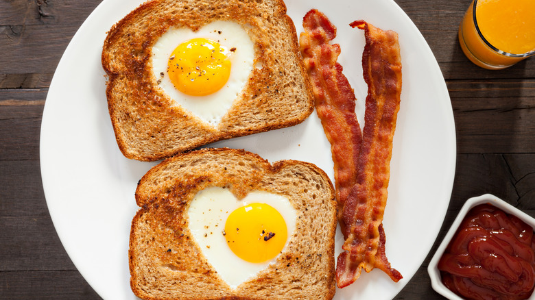 An overhead close up shot of heart-shaped eggs inside toast, a couple of bacon strips and fresh squeezed orange juice and ketchup