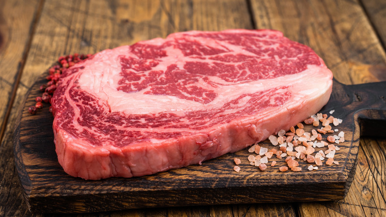 A raw ribeye steak sits on a wooden cutting board surrounded by rock salt