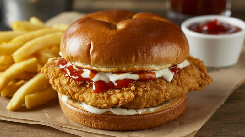 A breaded chicken sandwich on a bun with mayo and ketchup next to french fries.