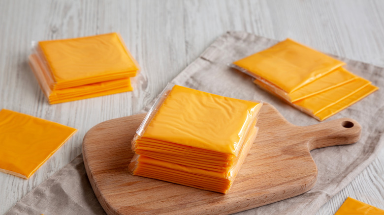 Slices of plastic-wrapped American cheese on and around wood cutting board.