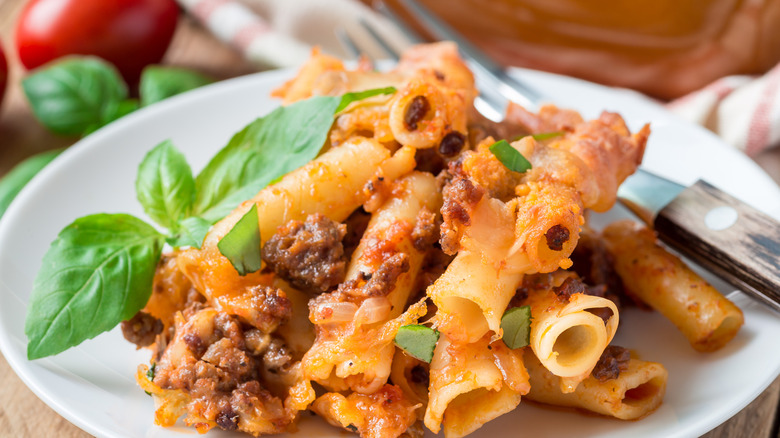 small portion of insufficiently sauced baked ziti on a white plate with a cluster of whole basil leaves and a fork