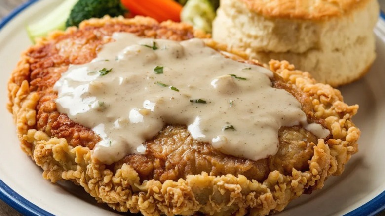 Close-up of chicken fried steak with cream gravy on a plate with a biscuit and vegetables.