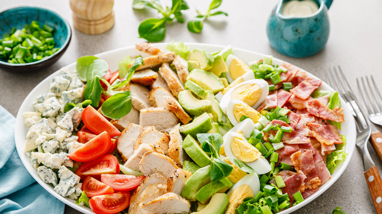 Cobb salad in a white bowl on a table with utensils and a pitcher of dressing