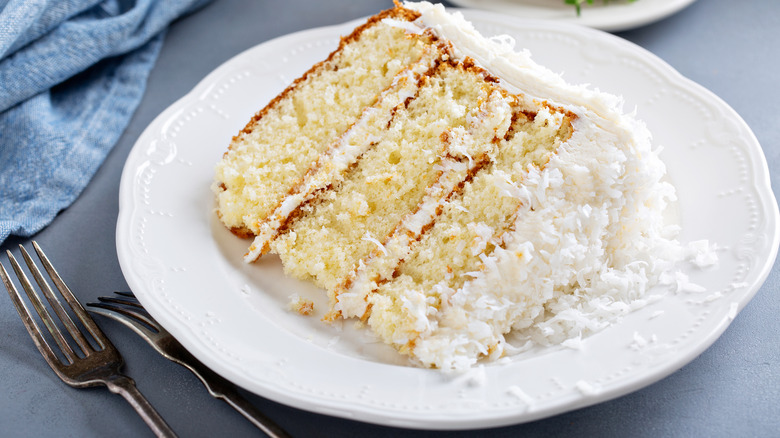 slice of coconut layer cake on a white plate next to a crumpled blue cloth napkin and two forks