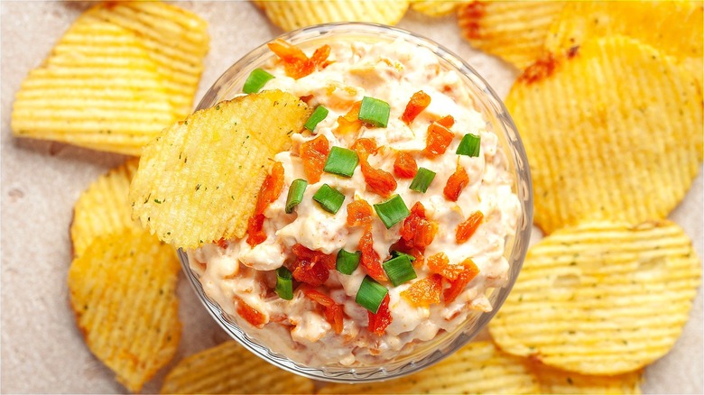 French onion dip in a glass bowl topped with chopped green onions, bacon bits, and served with ridged potato chips