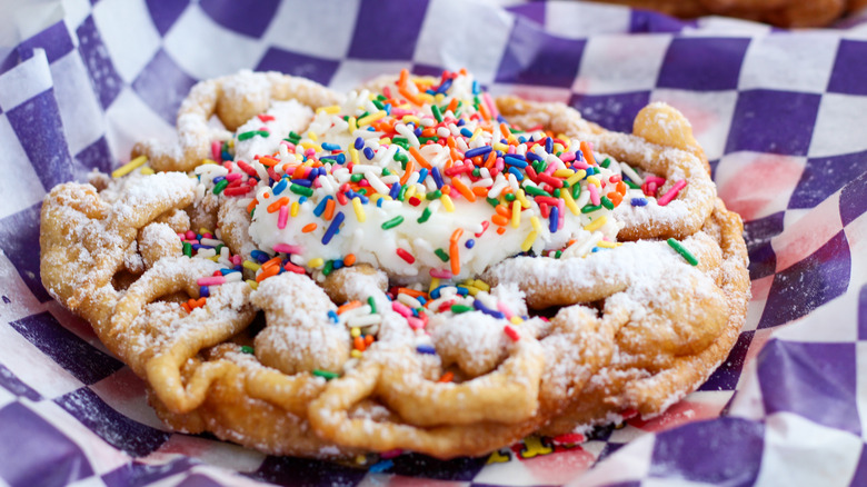 Funnel cake on served on blue and white checkered paper topped with powdered sugar, icing, and sprinkles