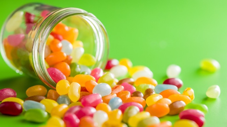 Different colors of jelly beans spilling out of an overturned mason jar on a green surface.