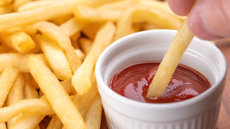 A ramekin of ketchup with a french fry being dipped into it.