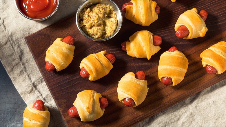 Evenly spaced pigs in a blanket on a dark wood serving board near small bowls of ketchup and grainy mustard