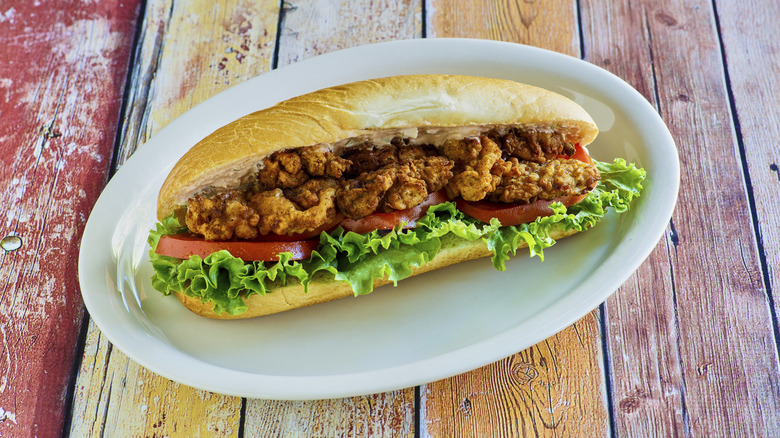 fried oyster po'boy with lettuce and tomato on an oval white plate sitting on a weathered wooden picnic table