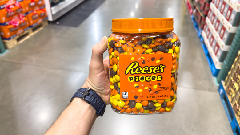 Person holding large container of Reese's Pieces candy inside grocery store.
