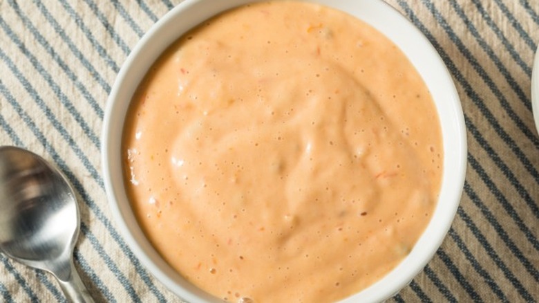 A white ramekin filled with thousand island dressing sitting on a striped placemat next to a spoon.