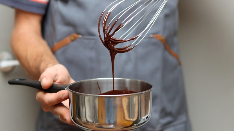 A man in an apron whisking chocolate ganache in a saucepan.
