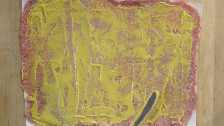 Mustard spread on ground chuck flattened on parchment paper over wooden surface