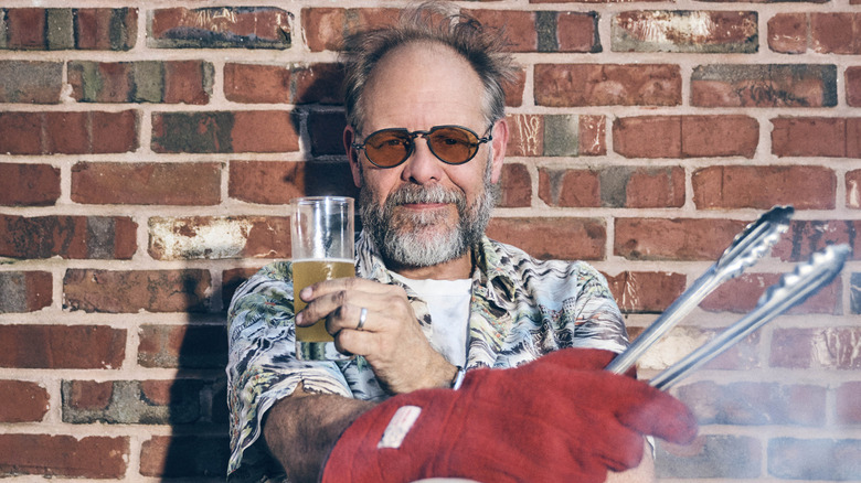 Alton Brown holds tongs and a beer while leaning against a brick wall.