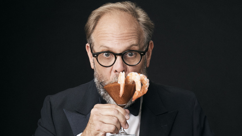 Alton Brown wears a tuxedo and is holding up a shrimp cocktail.