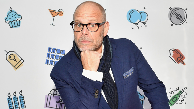 Alton Brown wearing a suit and scarf resting his head on his fist.