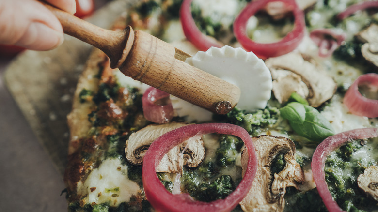 A pizza cutter slices through a pie topped with onion rings and mushrooms