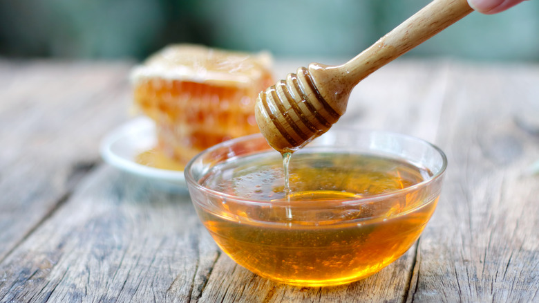 Honey being drizzled into a bowl with a honey dipper with honeycomb in the background