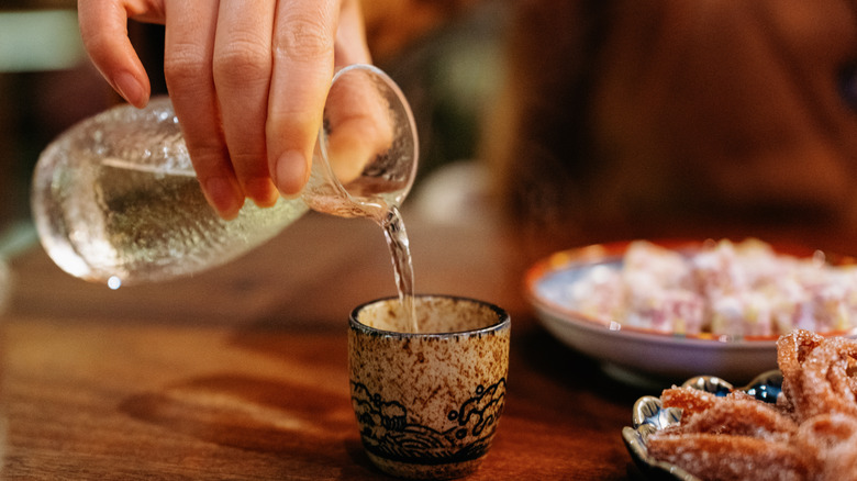 Close up of a hand pouring sake into a small cup with food nearby, all on a wooden table.