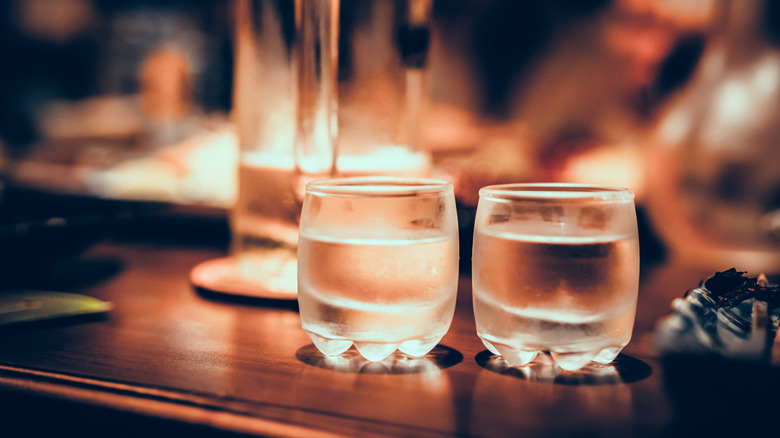 Close up of two glasses of sake on a wooden table with items blurred in the background.