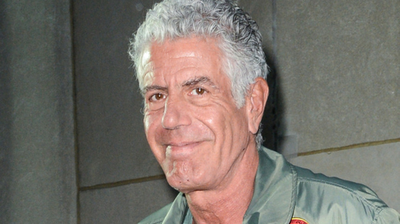 Anthony Bourdain smiling as he walks outside in a green bomber jacket