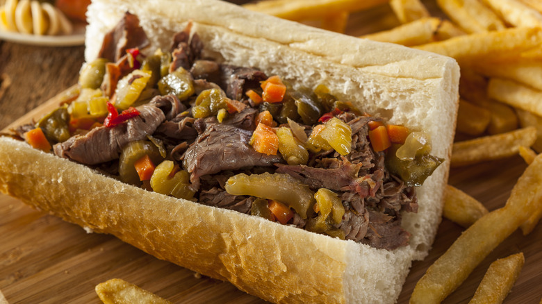 An Italian Beef sandwich next to loose French fries on a wooden board.